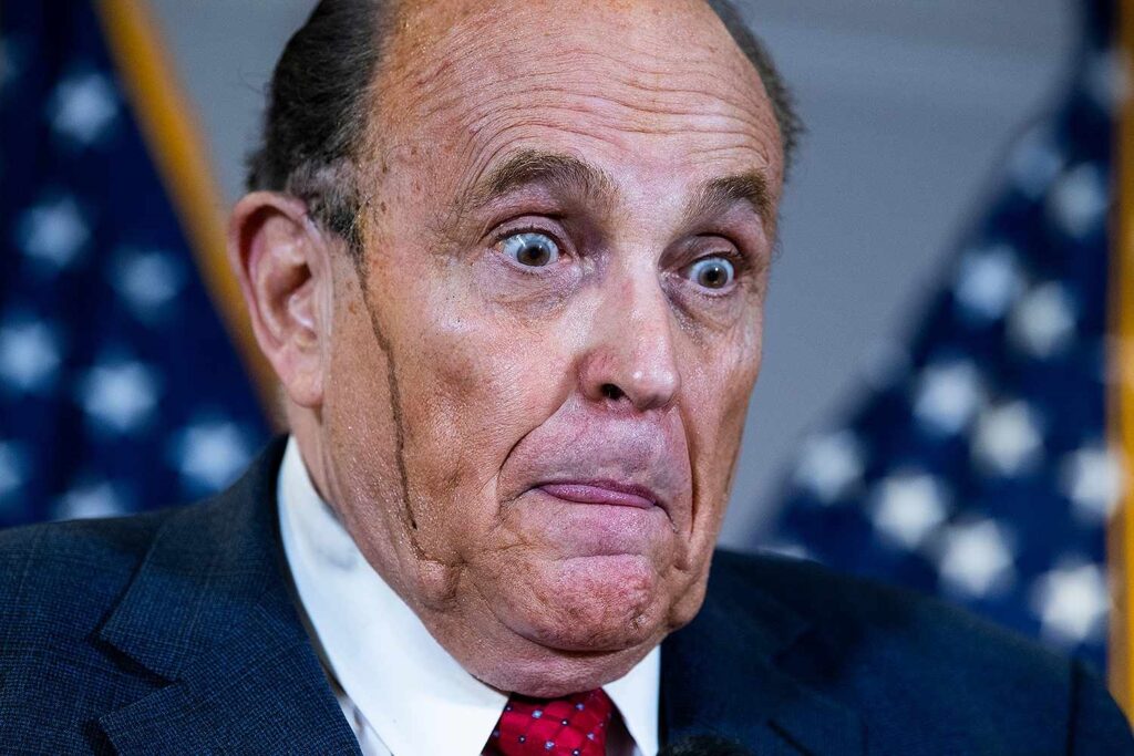 Imagen de Rudy Giuliani, abogado de TRUMP!, ex-Alcalde de Nueva York y tipo particular, que en esta imagen está poniendo una cara rara, sacando la punta de la lengua mientras abre mucho los ajos, ya la vez le está corriendo un churretón de color negro, probablemente del tinte del pelo, por toda la mejilla derecha.