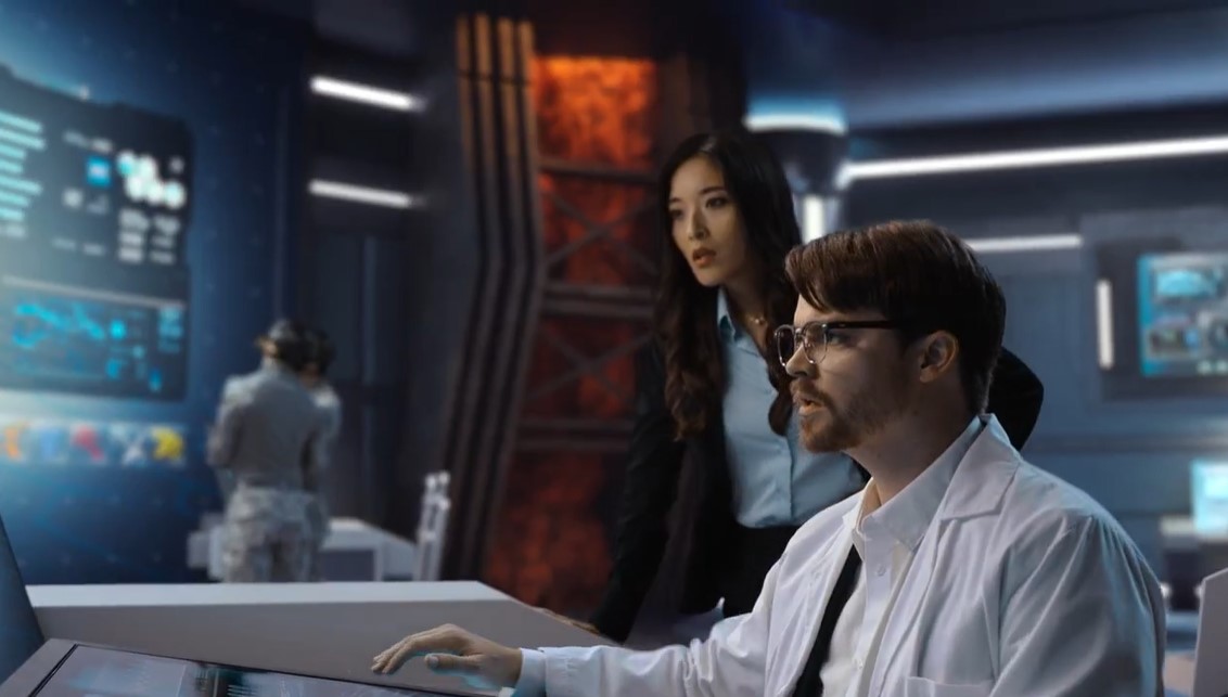 Imagen en la que vemos a un tipo vestido de científico sentado a una mesa, una mujer de traje está tras él, todo el espacio parece estar diseñado en 3D. Por detrás.