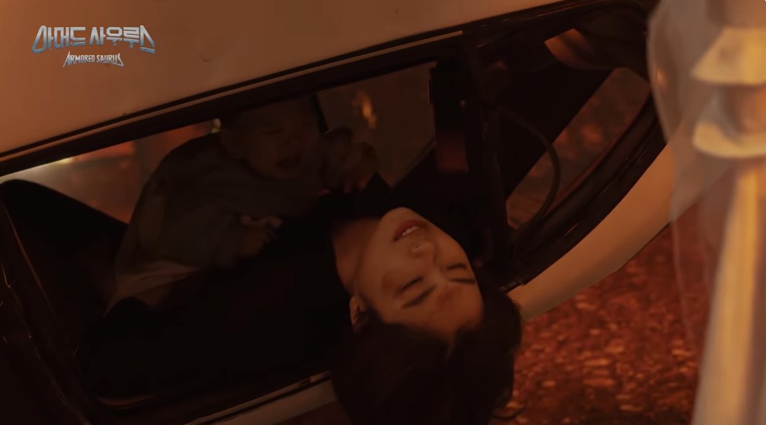 Imagen en la que vemos un coche bocabajo, hay alguien a la derecha, vestida de blanco y con tules o algo. De la ventanilla vemos una mujer saliendo, con un bebé encima.