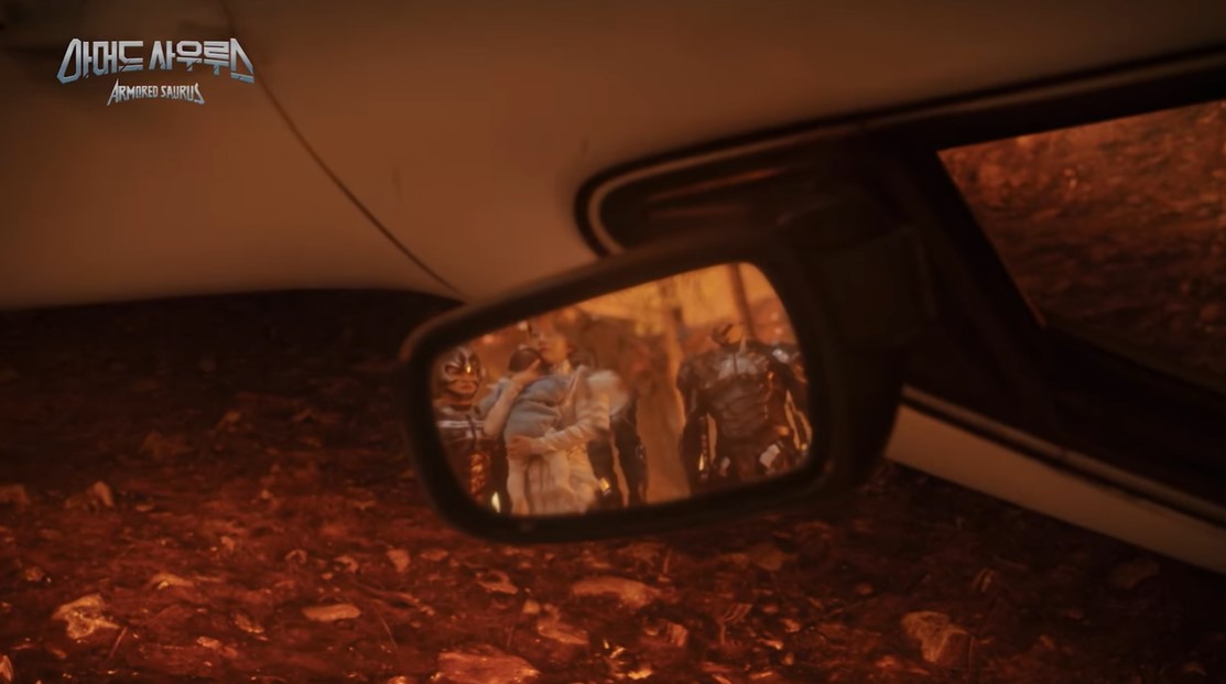 Imagen del coche bocabajo, se centra en un retrovisor en el que vemos a una mujer de blanco, junto a varios tipos con armaduras raras, la señora lleva al bebé en brazos.